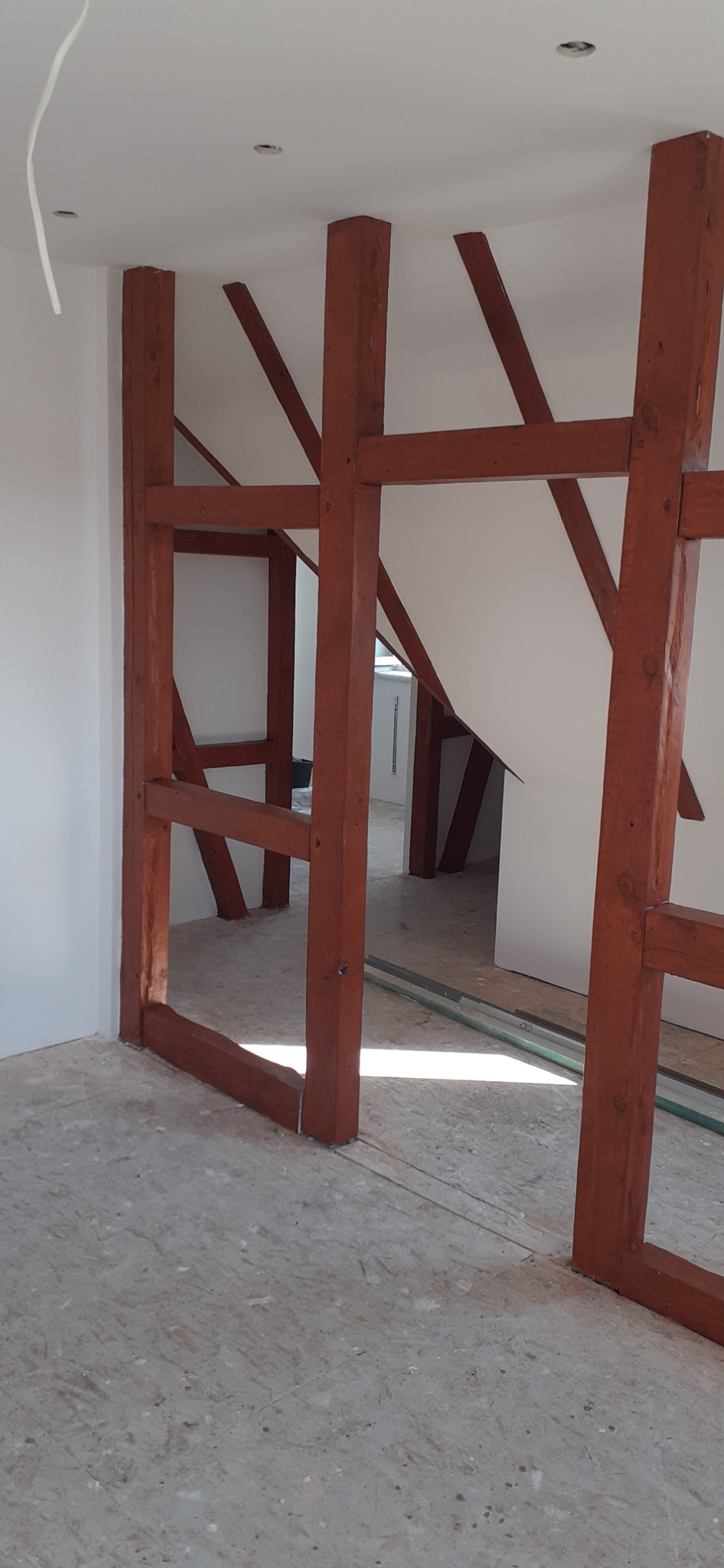 Konstrukcja szkieletowa z belek drewnianych w odcieniach brązu, tworząca ramę wewnątrz pomieszczenia, widoczne fragmenty białych ścian i sufitu, surowa podłoga z płyty OSB, w tle widok na kolejne...