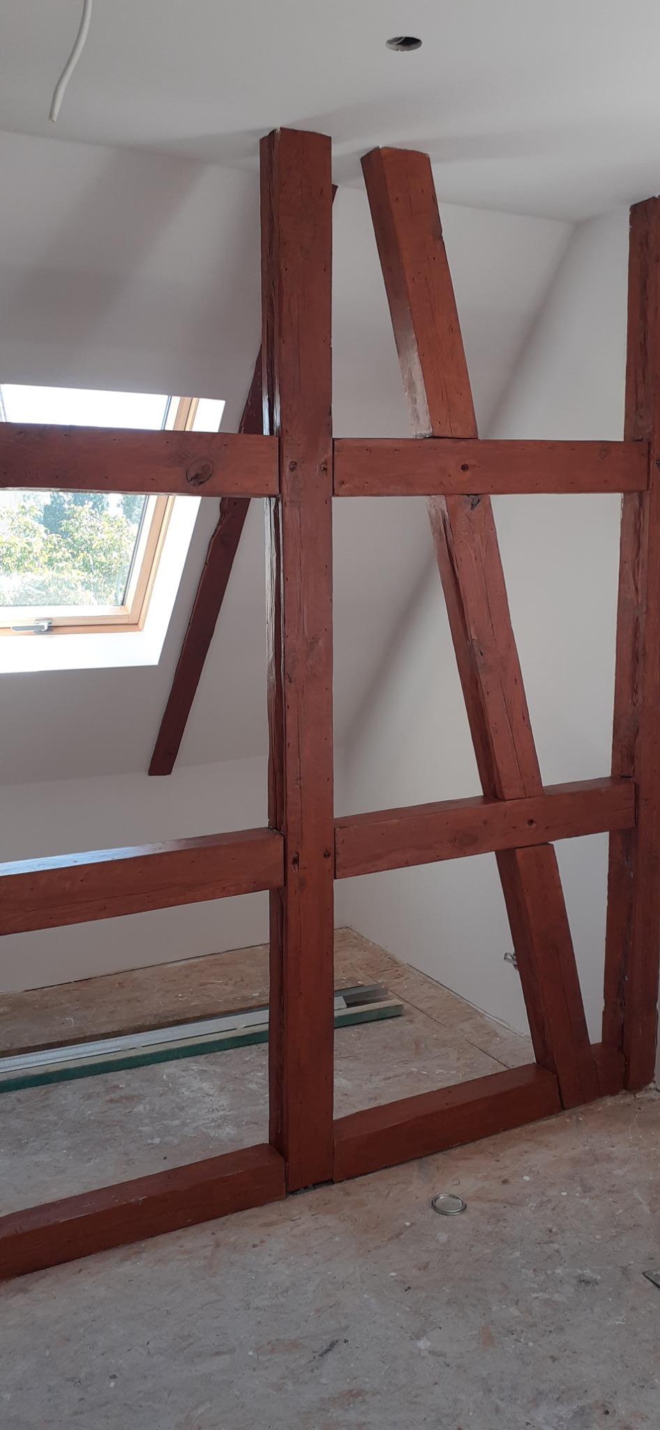 Fragment konstrukcji szkieletowej z belek w kolorze czerwono-brązowym, widok na poddasze z oknem dachowym, surowy stan podłogi z widocznymi listwami przypodłogowymi.