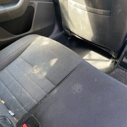 Brak - Bardzo zabrudzone, szare, tekstylne siedzenie samochodu z widocznymi plamami i okruchami, z czerwonym zatrzaskiem pasów bezpieczeństwa.