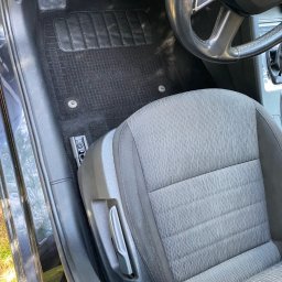 Brak - Wnętrze samochodu z czarnym dywanikiem podłogowym o fakturze kratki i szarą tapicerką siedzenia kierowcy, widoczna kierownica i fragment deski rozdzielczej.