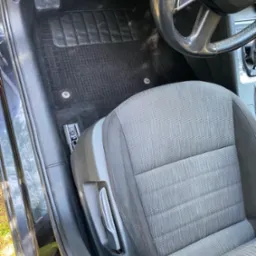 Wnętrze samochodu z czarnym dywanikiem podłogowym o fakturze kratki i szarą tapicerką siedzenia kierowcy, widoczna kierownica i fragment deski rozdzielczej.