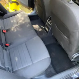 Wnętrze samochodu z wyczyszczoną tylną kanapą i dywanikami, widoczny odkurzacz piorący w tle, czarne obicia foteli i dywaników.