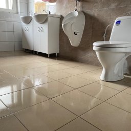 Brak - Widok z dołu na umywalki z szafkami, pisuar i toaletę w jasnej łazience wyłożonej beżowymi płytkami, z odbiciami na podłodze.