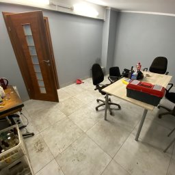 Brak - Pokój po remoncie z otwartymi drzwiami, biurkiem z narzędziami i pozostałościami po pracy, widoczne plamy na podłodze i puste butelki w skrzynkach.
