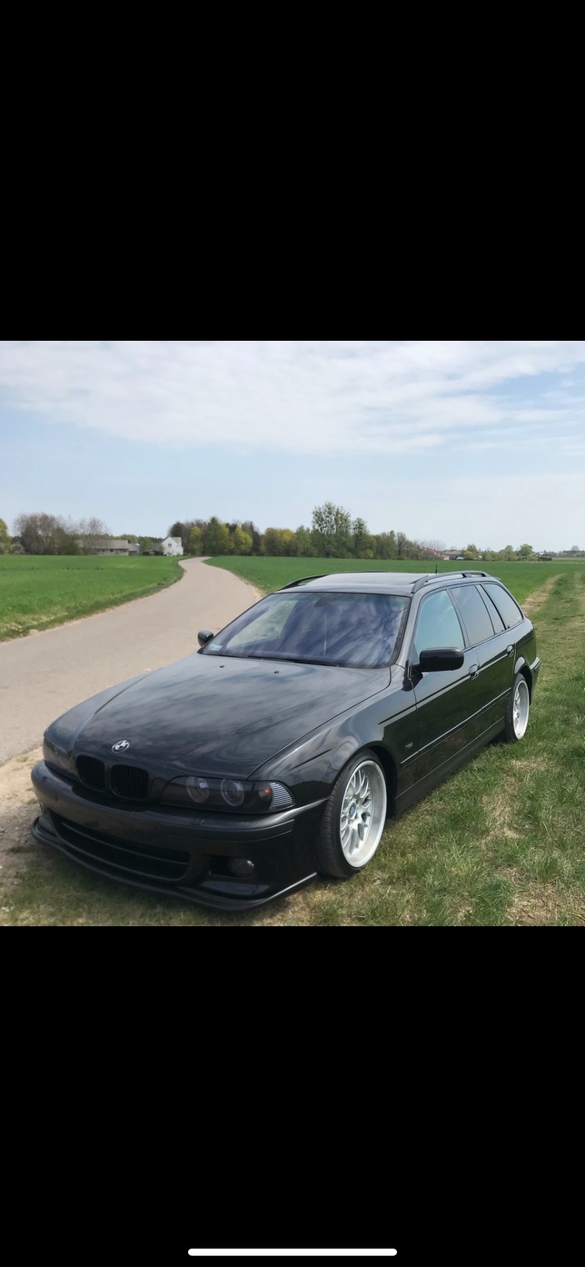 Czarny samochód kombi BMW serii 5 E39 stoi na trawie obok asfaltowej drogi, w tle budynki gospodarcze i drzewa.