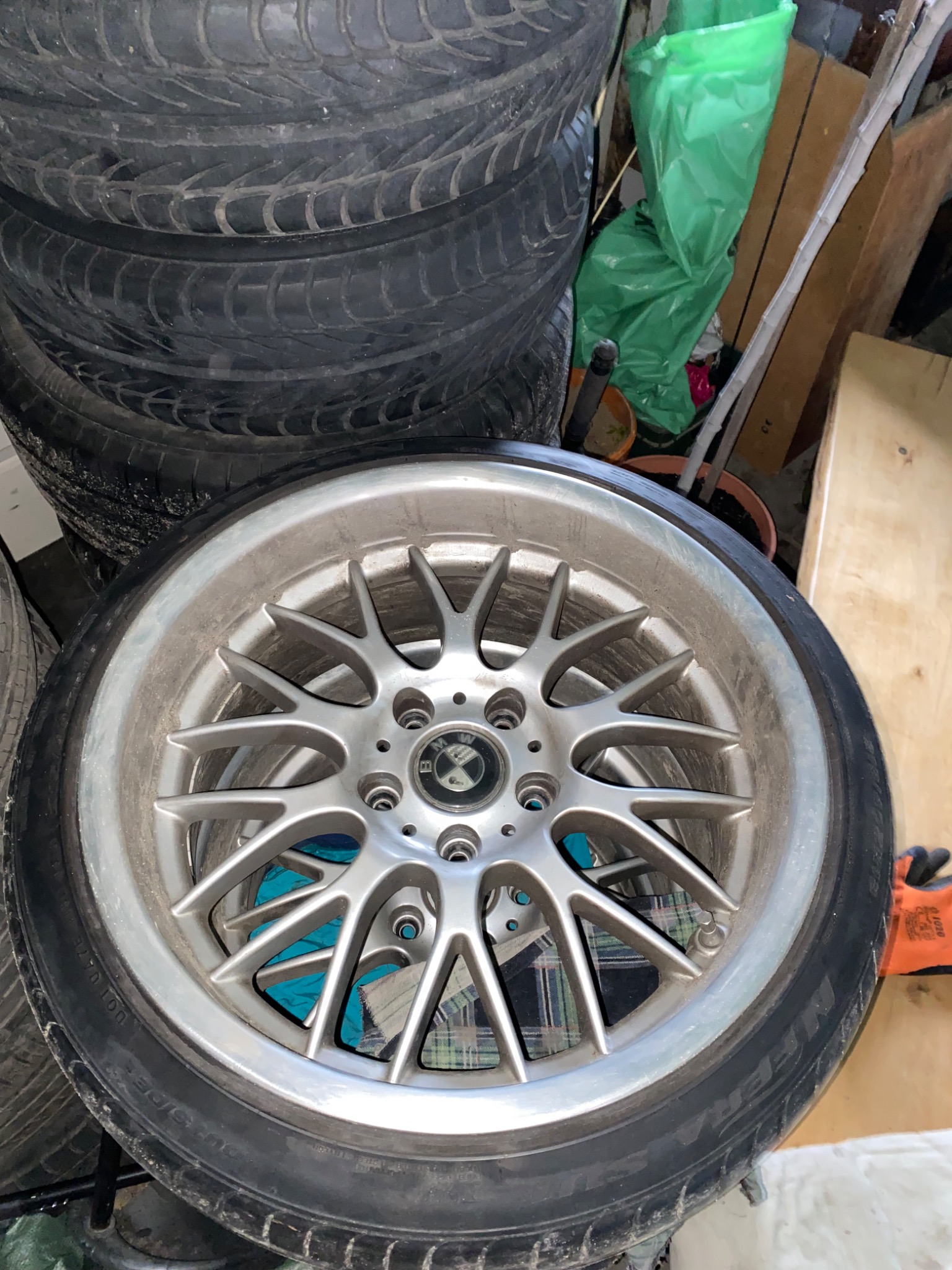 Stos opon samochodowych z aluminiową felgą o szprychowym wzorze, widoczne ślady użytkowania i zabrudzenia, w tle nieuporządkowane przedmioty w garażu.