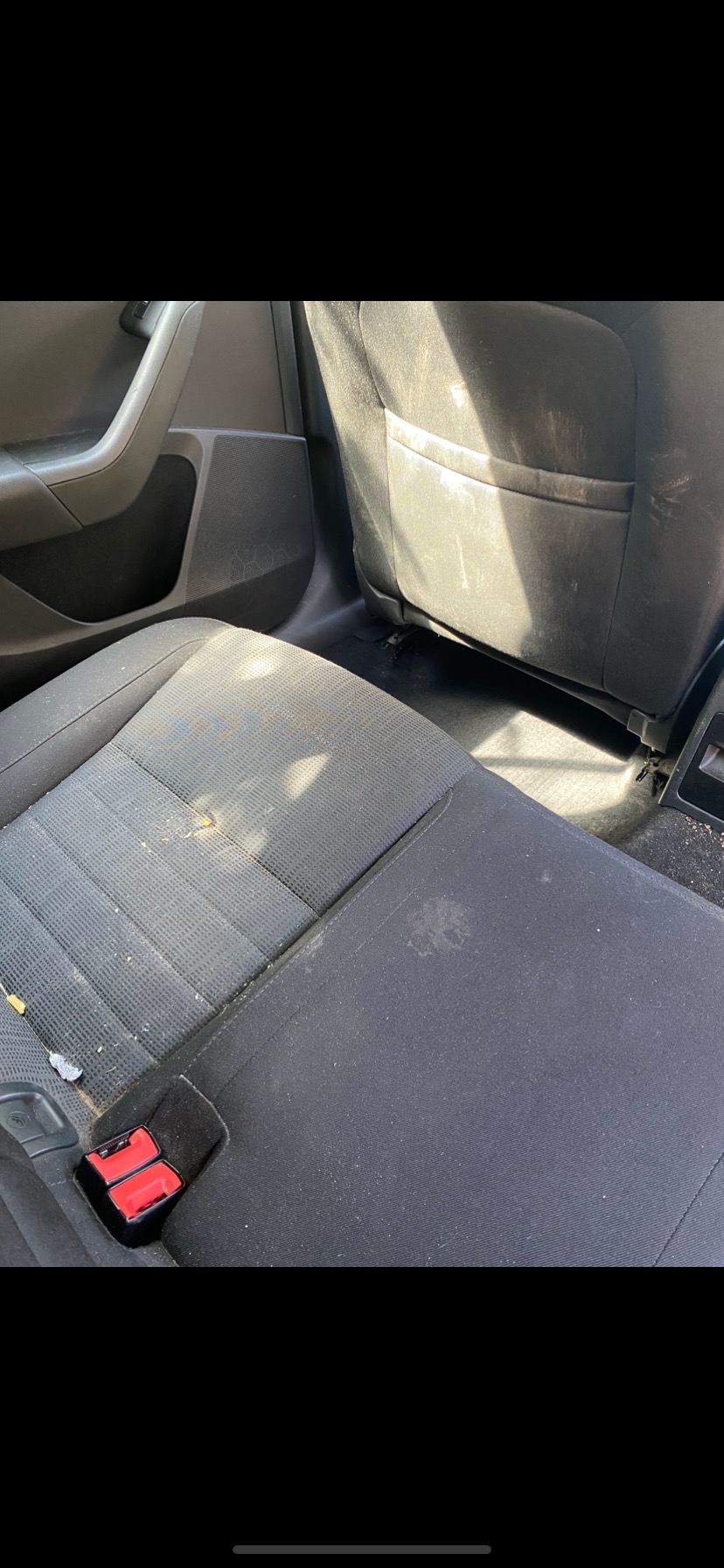 Bardzo zabrudzone, szare, tekstylne siedzenie samochodu z widocznymi plamami i okruchami, z czerwonym zatrzaskiem pasów bezpieczeństwa.