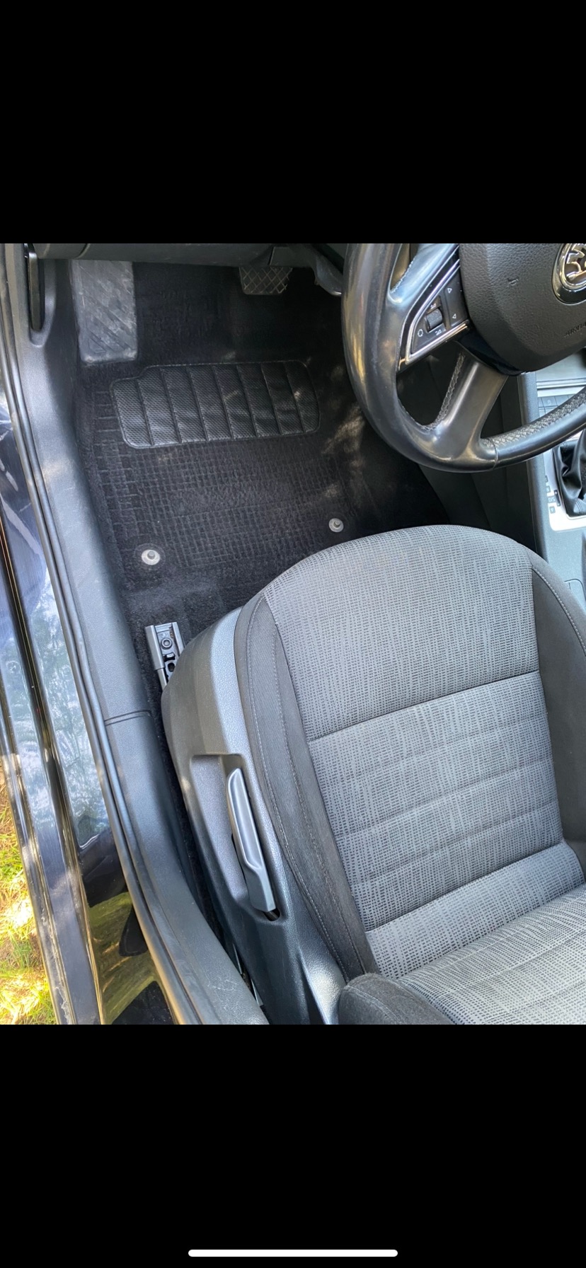 Wnętrze samochodu z czarnym dywanikiem podłogowym o fakturze kratki i szarą tapicerką siedzenia kierowcy, widoczna kierownica i fragment deski rozdzielczej.