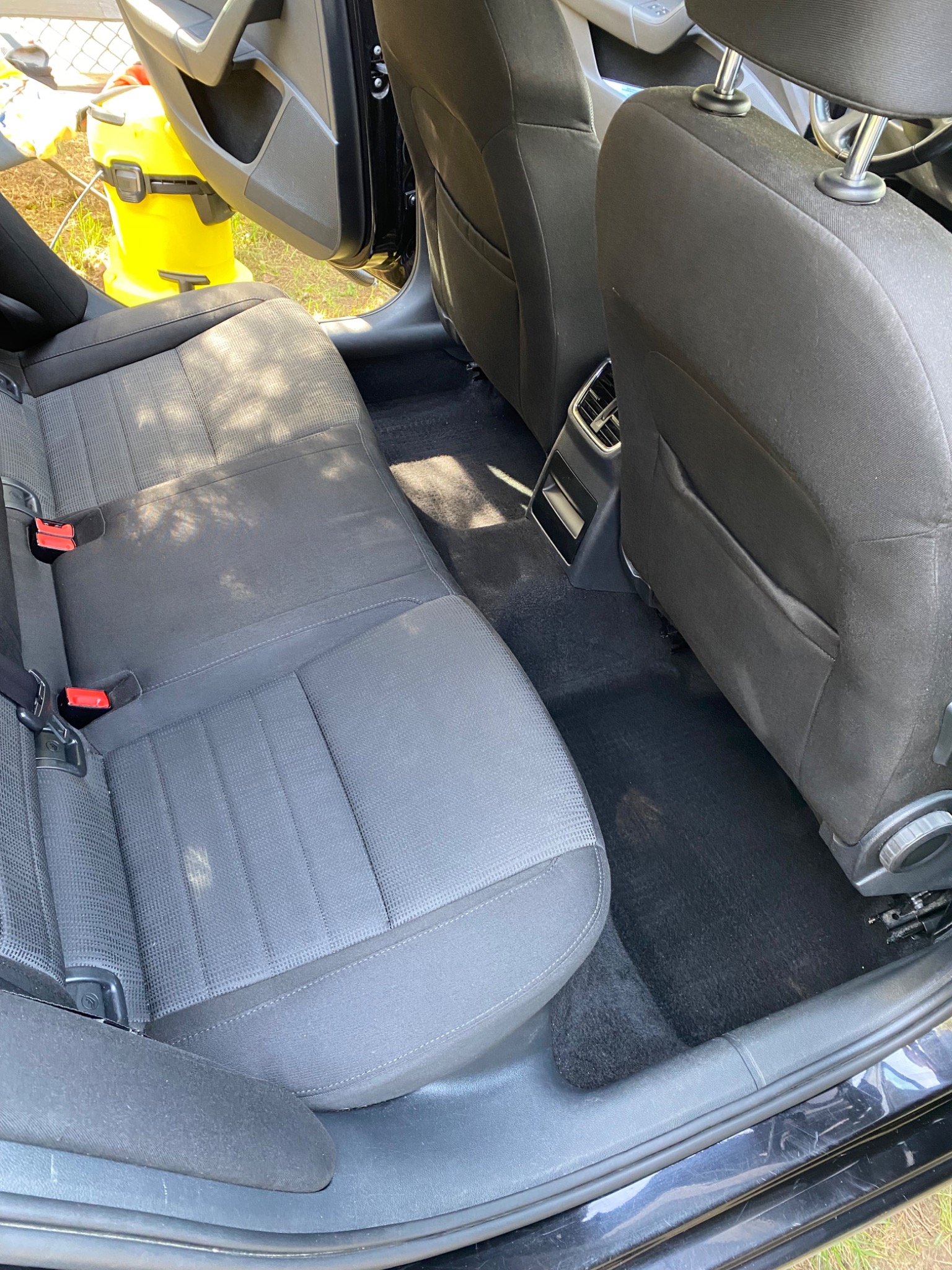 Wnętrze samochodu z wyczyszczoną tylną kanapą i dywanikami, widoczny odkurzacz piorący w tle, czarne obicia foteli i dywaników.