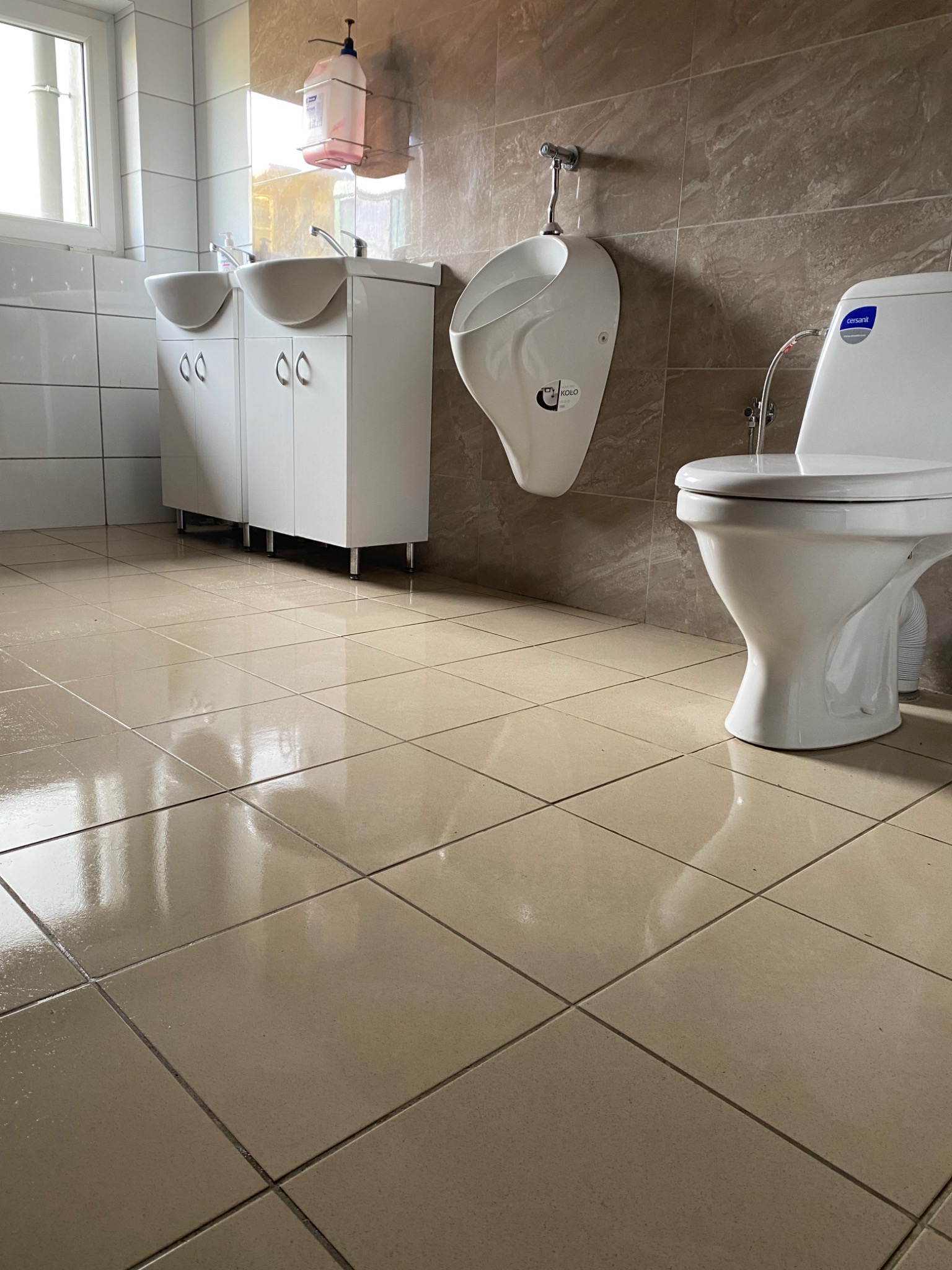 Widok z dołu na umywalki z szafkami, pisuar i toaletę w jasnej łazience wyłożonej beżowymi płytkami, z odbiciami na podłodze.