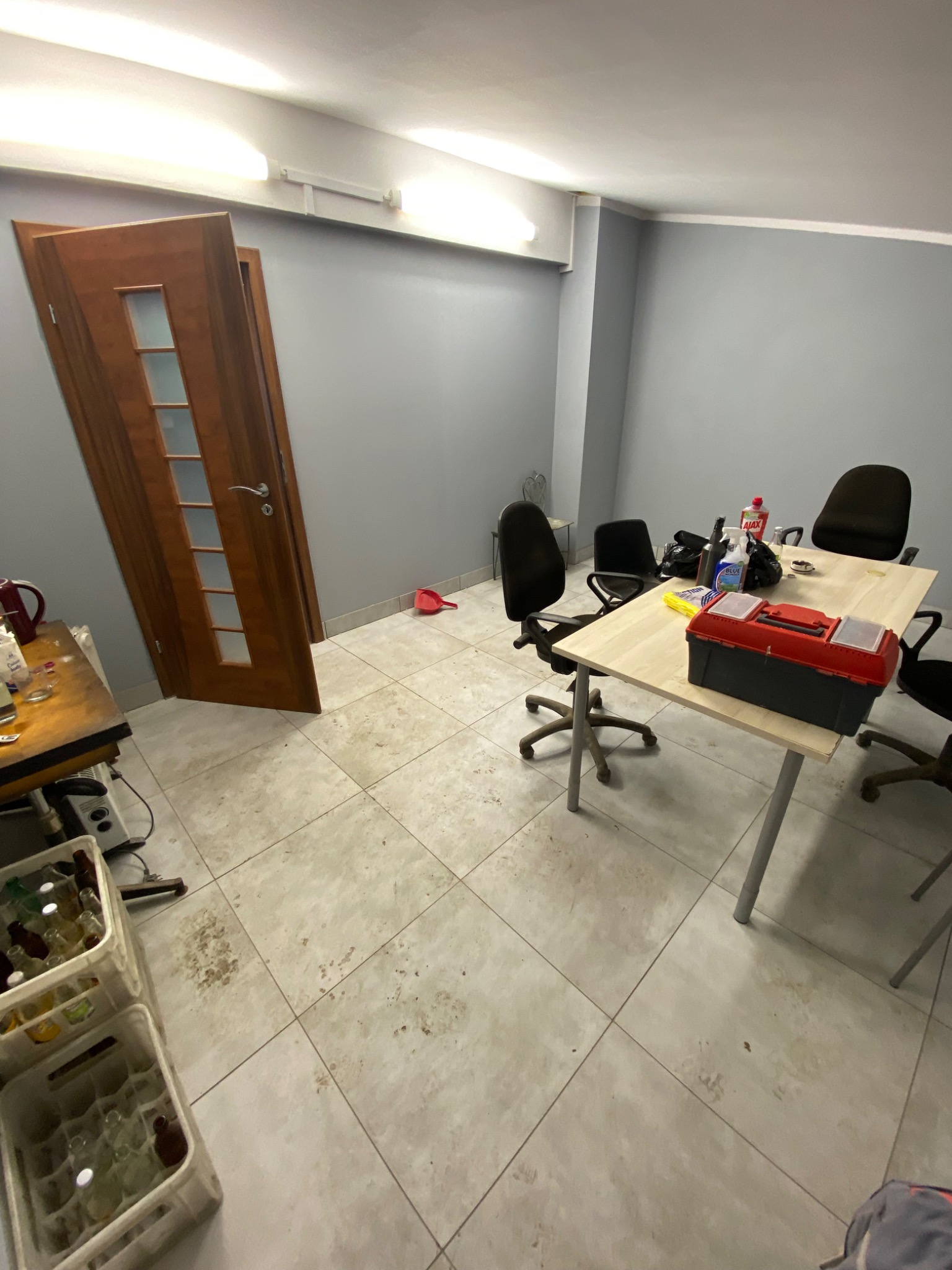 Pokój po remoncie z otwartymi drzwiami, biurkiem z narzędziami i pozostałościami po pracy, widoczne plamy na podłodze i puste butelki w skrzynkach.