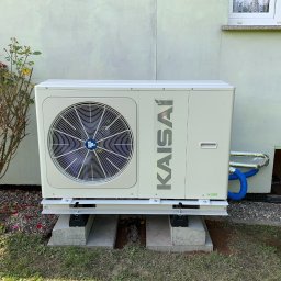Inżynieria Ciepła Sebastian Kazior - Zewnętrzna jednostka pompy ciepła marki Kaisai, zamontowana na betonowych bloczkach przed jasnym budynkiem z kwiatami w tle, widoczne niebieskie przewody.