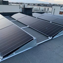 Inżynieria Ciepła Sebastian Kazior - Panele słoneczne zamontowane na płaskim dachu budynku mieszkalnego, widoczne elementy mocujące i okablowanie, tło stanowią budynki w oddali pod pochmurnym niebem.