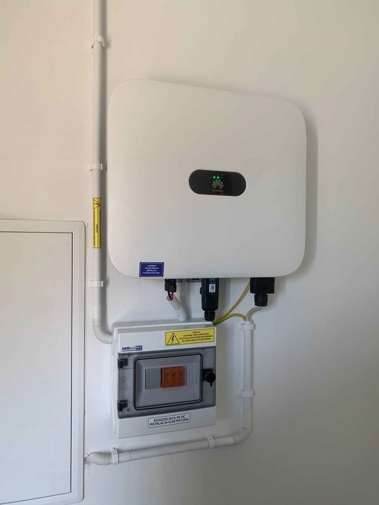 Biały falownik Huawei z trzema zielonymi diodami, zamontowany na białej ścianie nad rozdzielnicą PV DC z widoczną instalacją elektryczną i żółtą naklejką ostrzegawczą.