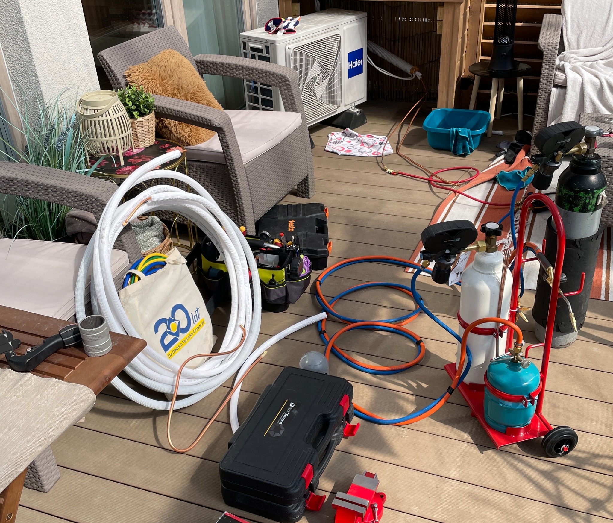 Instalacja klimatyzacji na tarasie: jednostka zewnętrzna Haier, zwój białych rur, butle z gazem na wózku, narzędzia w skrzynkach i torbie z logo 20 lat De Dietrich Technika Grzewcza.