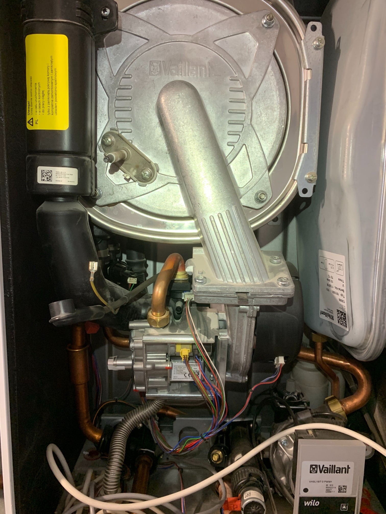 Wnętrze pieca gazowego marki Vaillant, widoczne miedziane rury, okablowanie, elementy sterujące i tabliczka znamionowa. Detal techniczny instalacji grzewczej.