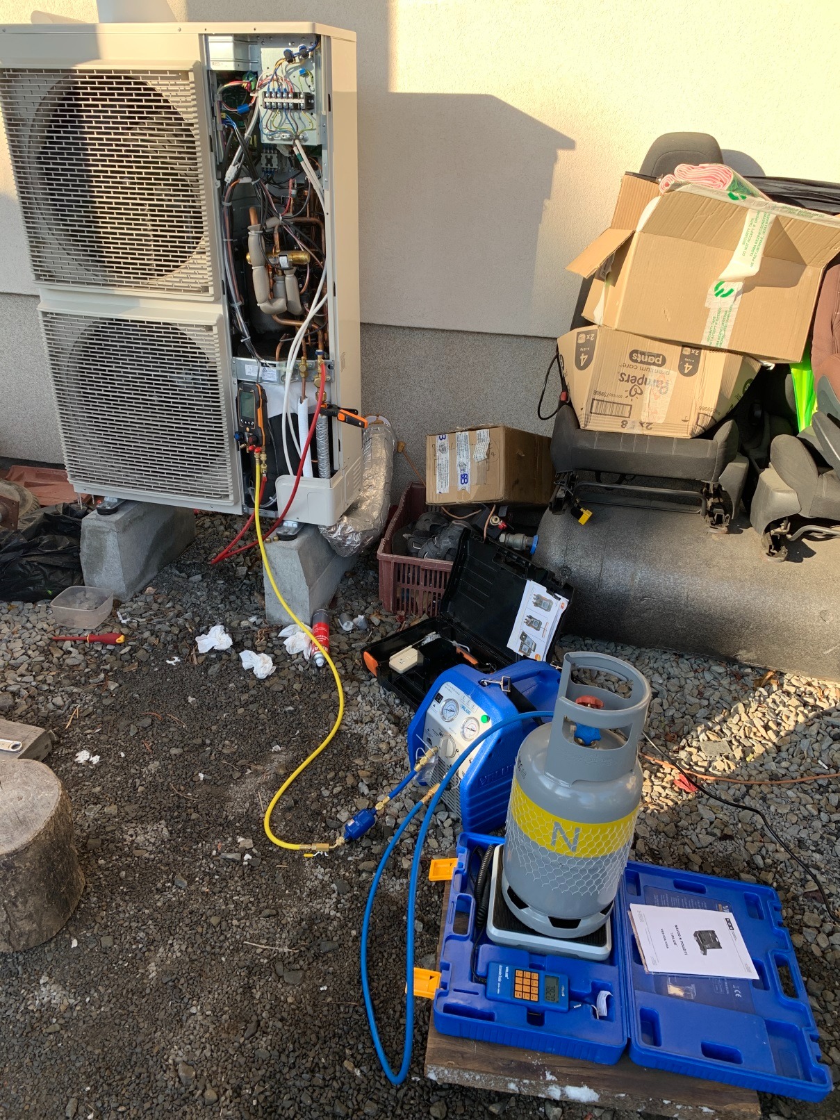 Otwarta obudowa zewnętrznej jednostki pompy ciepła podczas serwisu, widoczne podłączenia manometrów, butla z czynnikiem chłodniczym na wadze elektronicznej, narzędzia i kartony w tle.