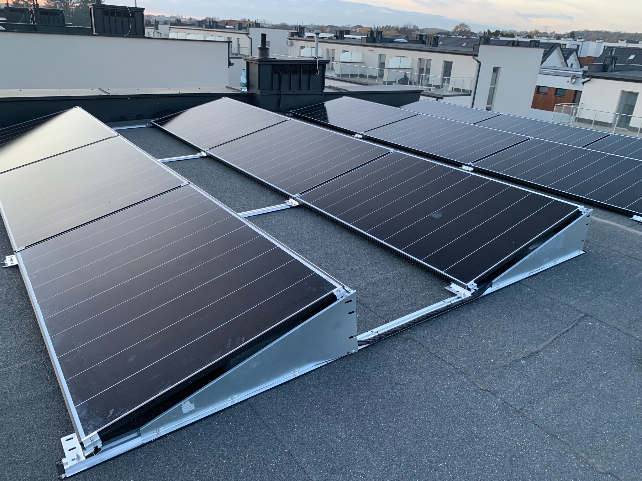Panele słoneczne zamontowane na płaskim dachu budynku mieszkalnego, widoczne elementy mocujące i okablowanie, tło stanowią budynki w oddali pod pochmurnym niebem.