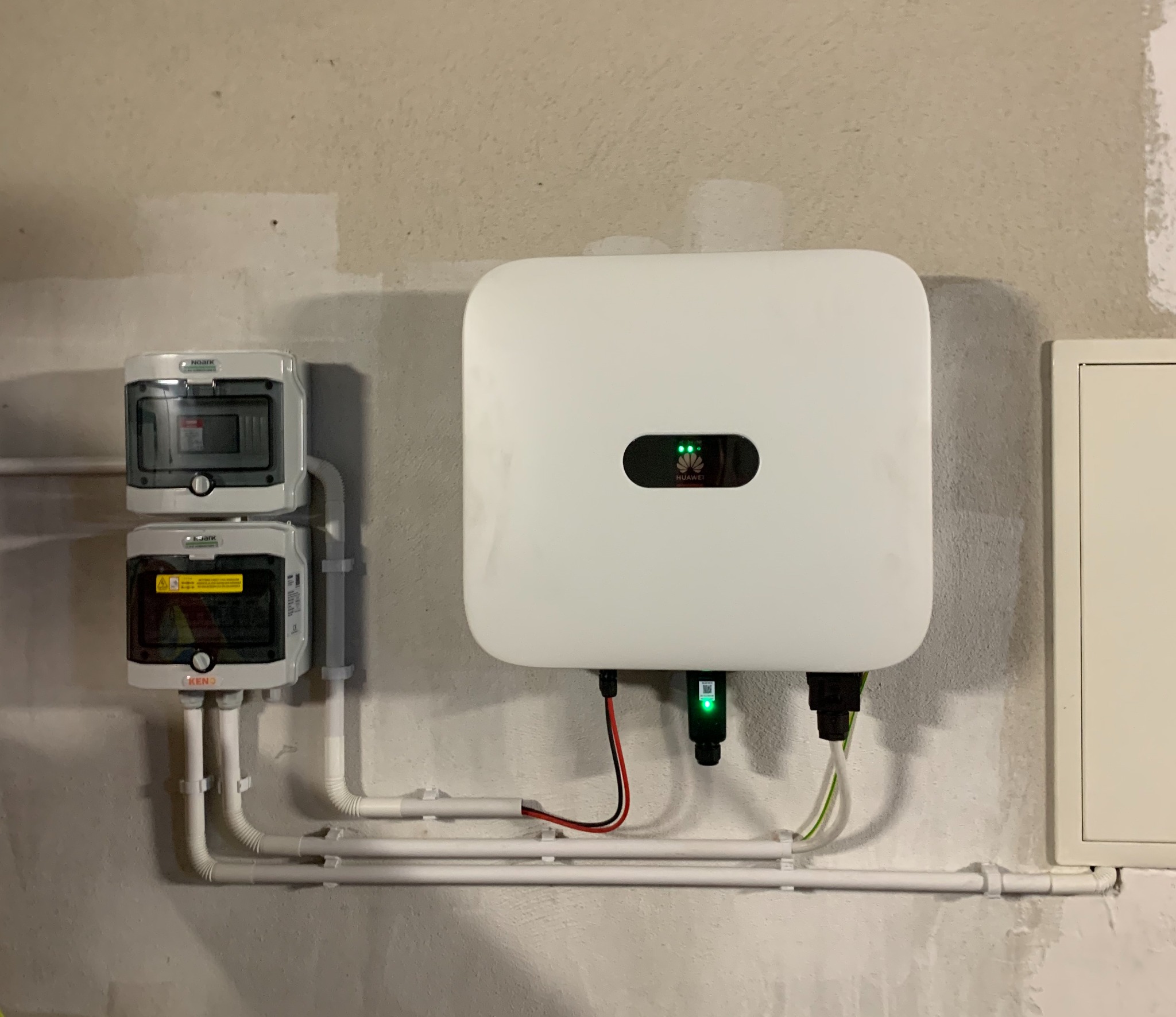 Biały inwerter fotowoltaiczny marki Huawei z zielonymi diodami sygnalizacyjnymi, zamontowany na szarej ścianie obok dwóch skrzynek elektrycznych Noark z widocznymi przewodami w białych rurkach PCV.