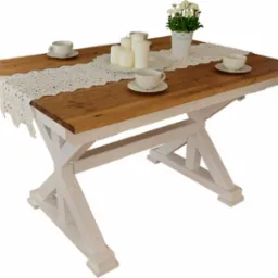 Drewniany stół z blatem w odcieniach brązu i bielonymi nogami w kształcie litery X, dekorowany koronkowym bieżnikiem, świecami, kwiatami w doniczce i filiżankami.