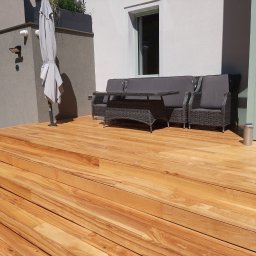 Michał Rychlicki Struga - Jasny, drewniany taras z meblami ogrodowymi z rattanu w odcieniach szarości, parasol i nowoczesny budynek w tle.