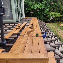 Michał Rychlicki Struga - Montaż desek na tarasie drewnianym, widoczne narzędzia, legary z tworzywa sztucznego i fragment budynku z dużymi oknami.