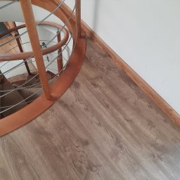 Michał Rychlicki Struga - Fragment drewnianych, kręconych schodów z metalowymi poręczami na podłodze pokrytej panelami laminowanymi o wzorze drewna, widok z góry.