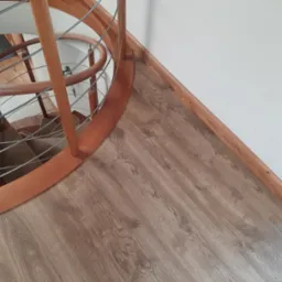 Fragment drewnianych, kręconych schodów z metalowymi poręczami na podłodze pokrytej panelami laminowanymi o wzorze drewna, widok z góry.