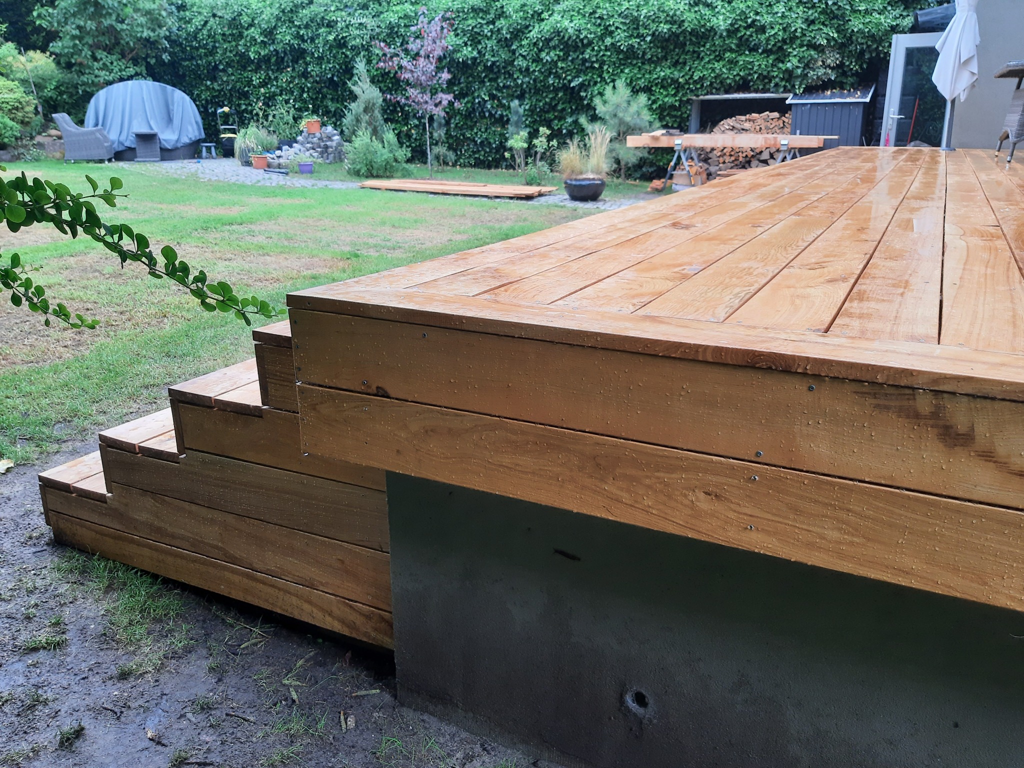 Nowo wybudowany, drewniany taras z niskimi schodami w ogrodzie, świeżo po deszczu, z widocznymi kroplami wody na deskach i elementami konstrukcyjnymi w tle.