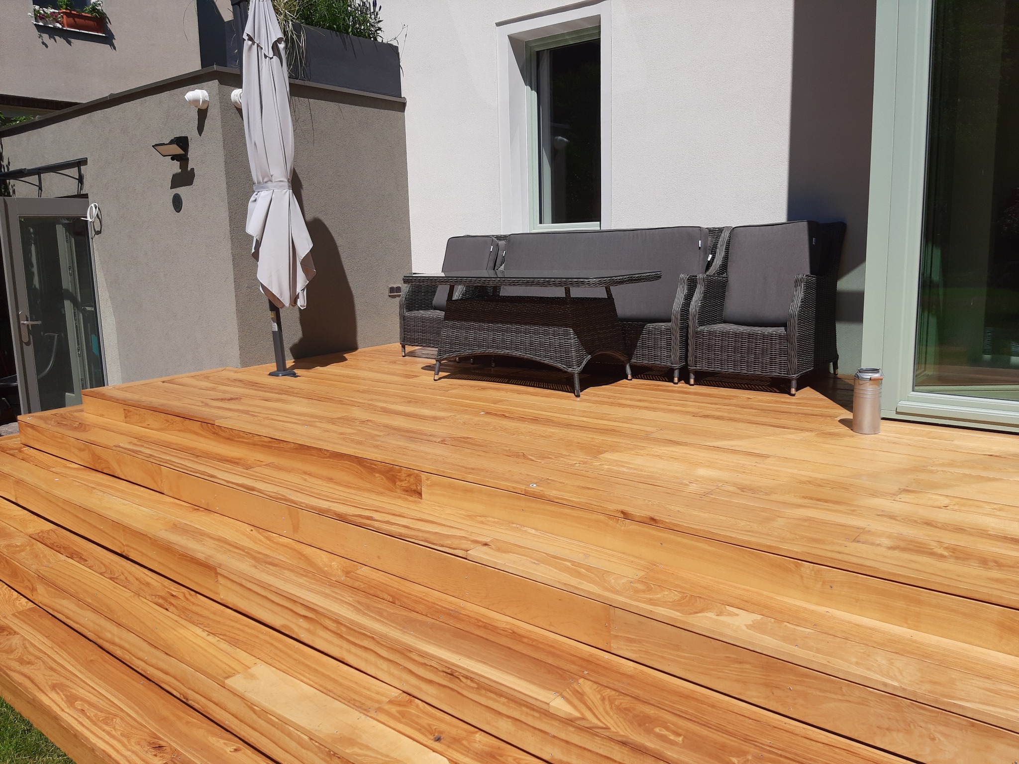 Jasny, drewniany taras z meblami ogrodowymi z rattanu w odcieniach szarości, parasol i nowoczesny budynek w tle.