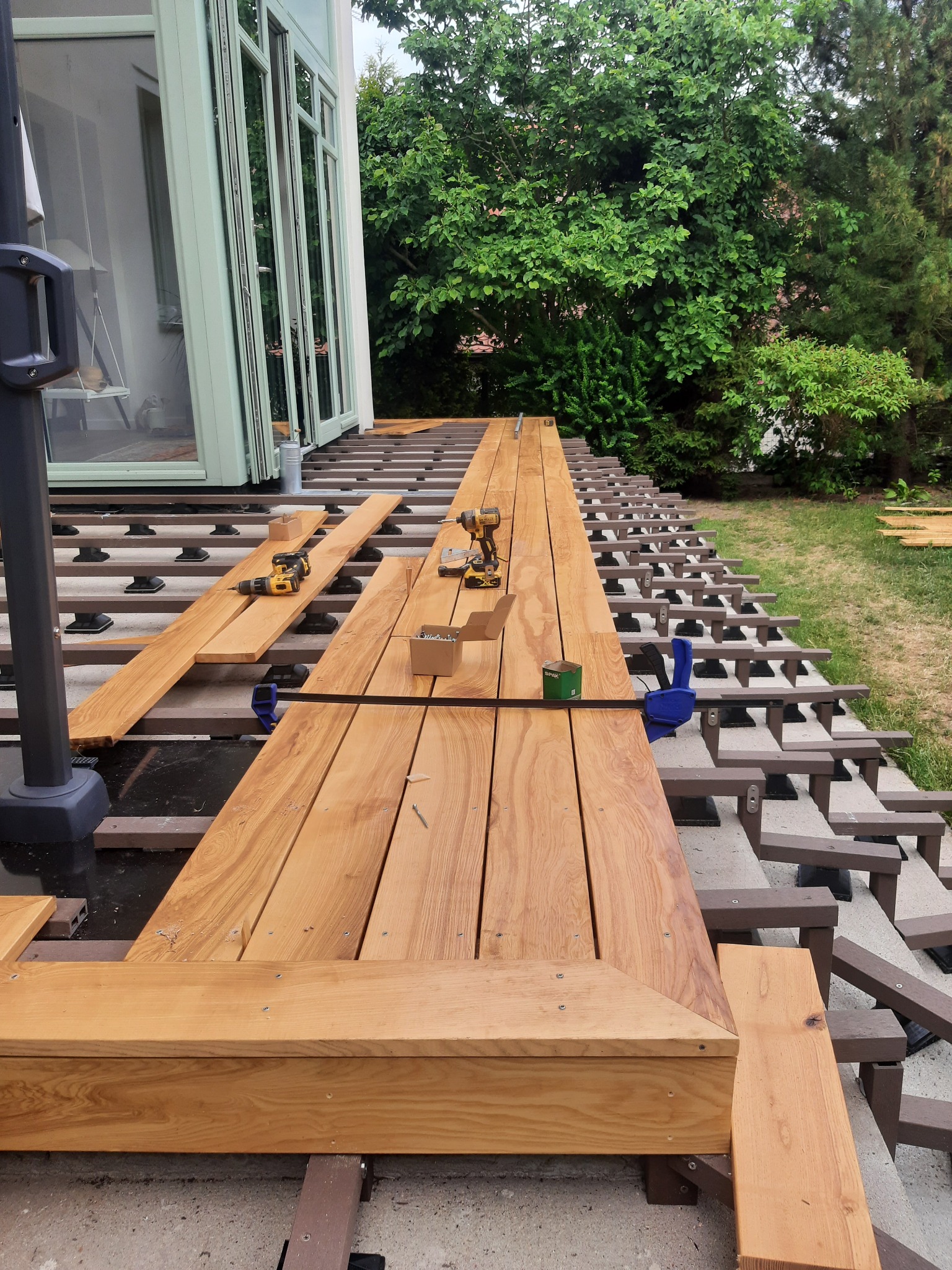 Montaż desek na tarasie drewnianym, widoczne narzędzia, legary z tworzywa sztucznego i fragment budynku z dużymi oknami.