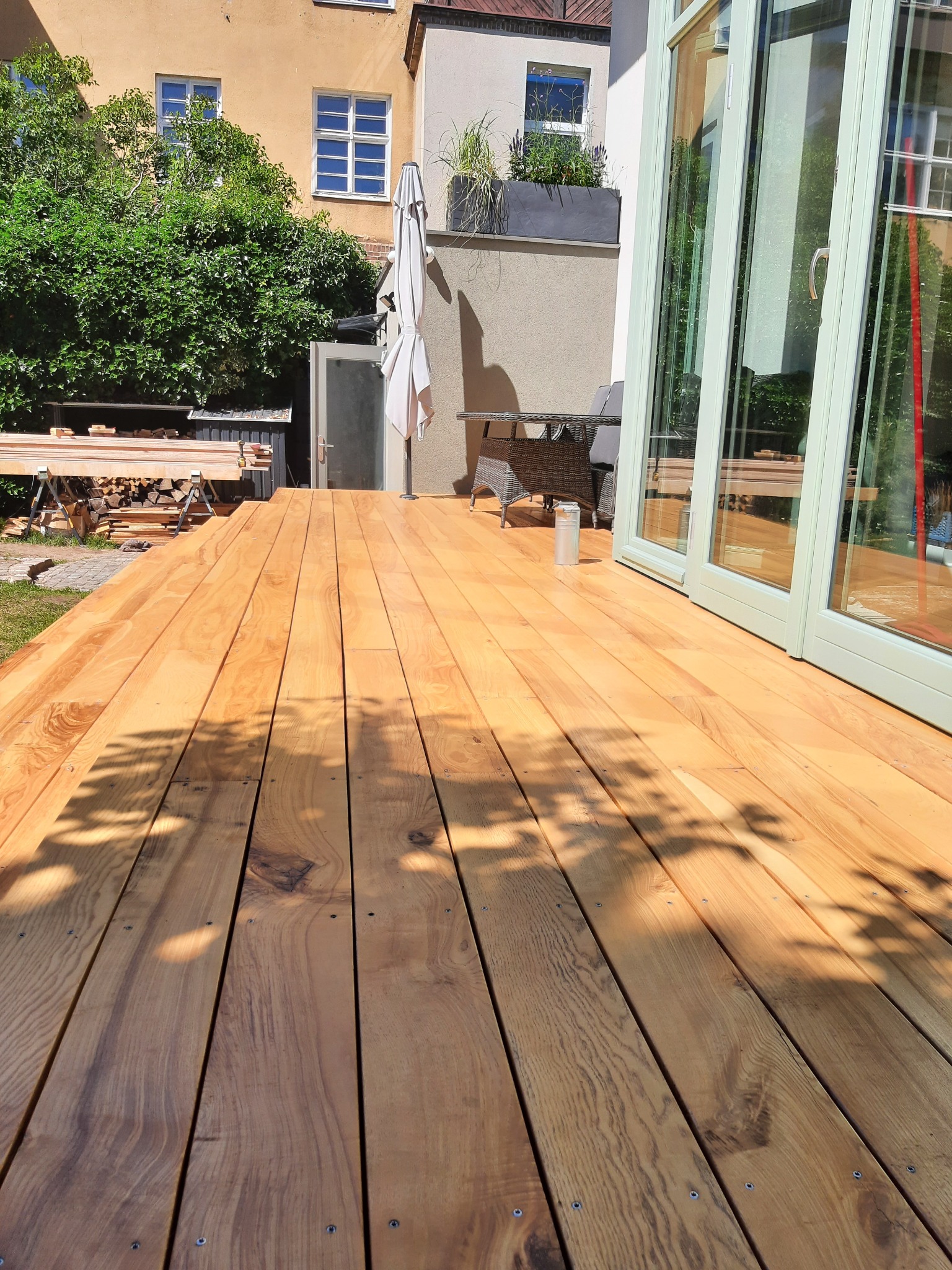 Świeżo ułożony taras z desek w ciepłym odcieniu drewna, z widocznymi śrubami mocującymi, przylegający do budynku z przeszklonymi drzwiami i elementami wyposażenia, z widocznymi narzędziami...