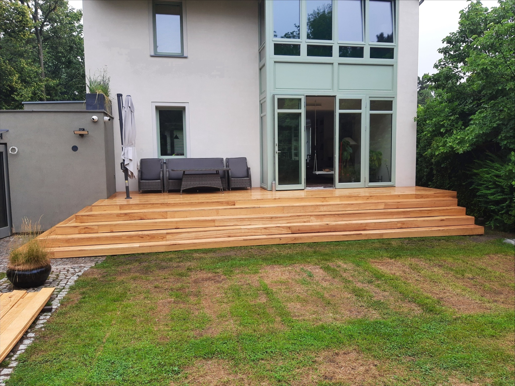 Nowoczesny taras drewniany z jasnego drewna, prowadzący do domu z dużymi oknami, uzupełniony o meble ogrodowe i dekoracyjną roślinność w donicy.