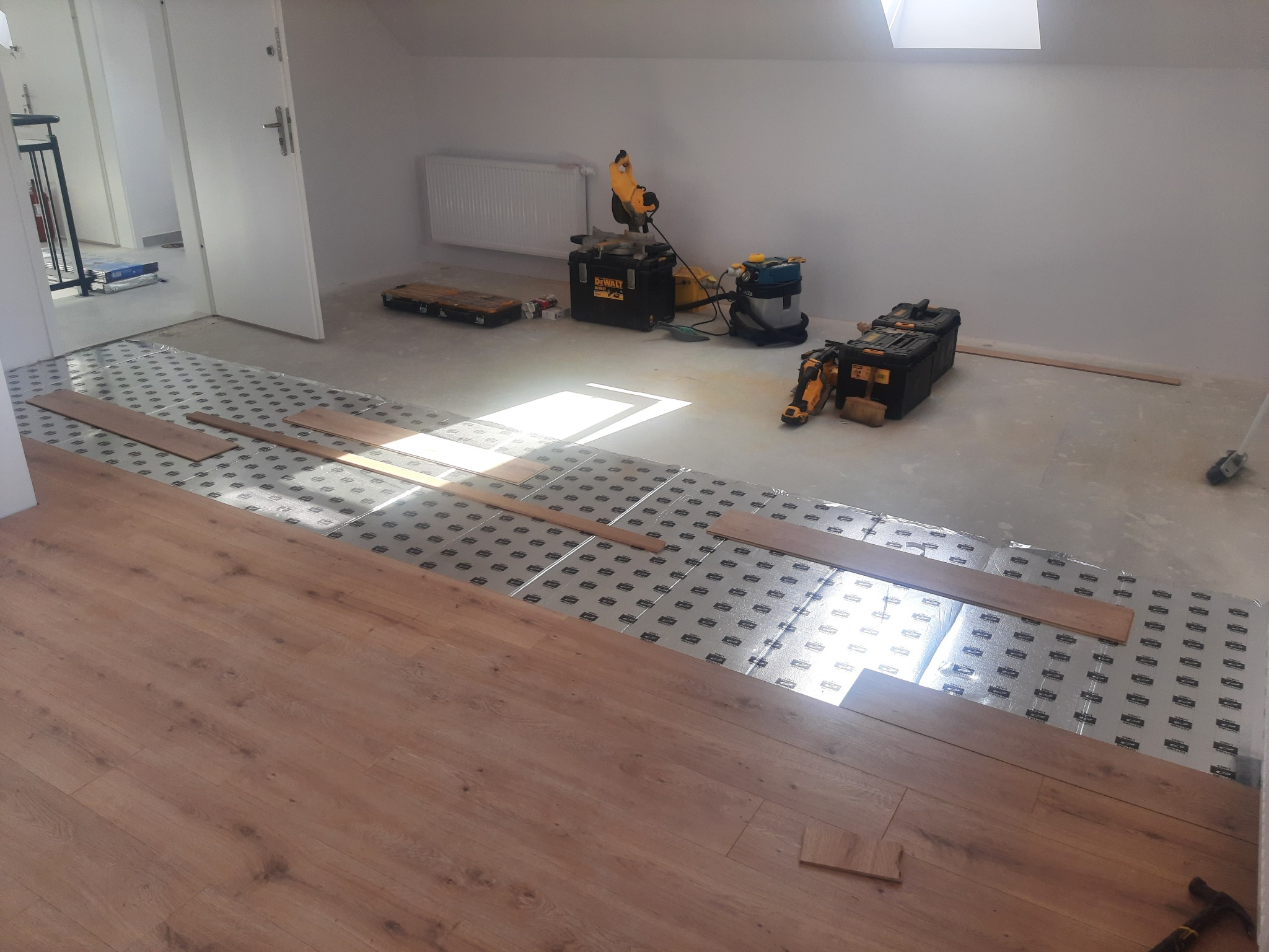 Montaż paneli laminowanych Egger w jasnym pokoju na poddaszu, widoczne narzędzia: piła ukośnica DeWalt, odkurzacz budowlany, skrzynki narzędziowe, podkład pod panele.