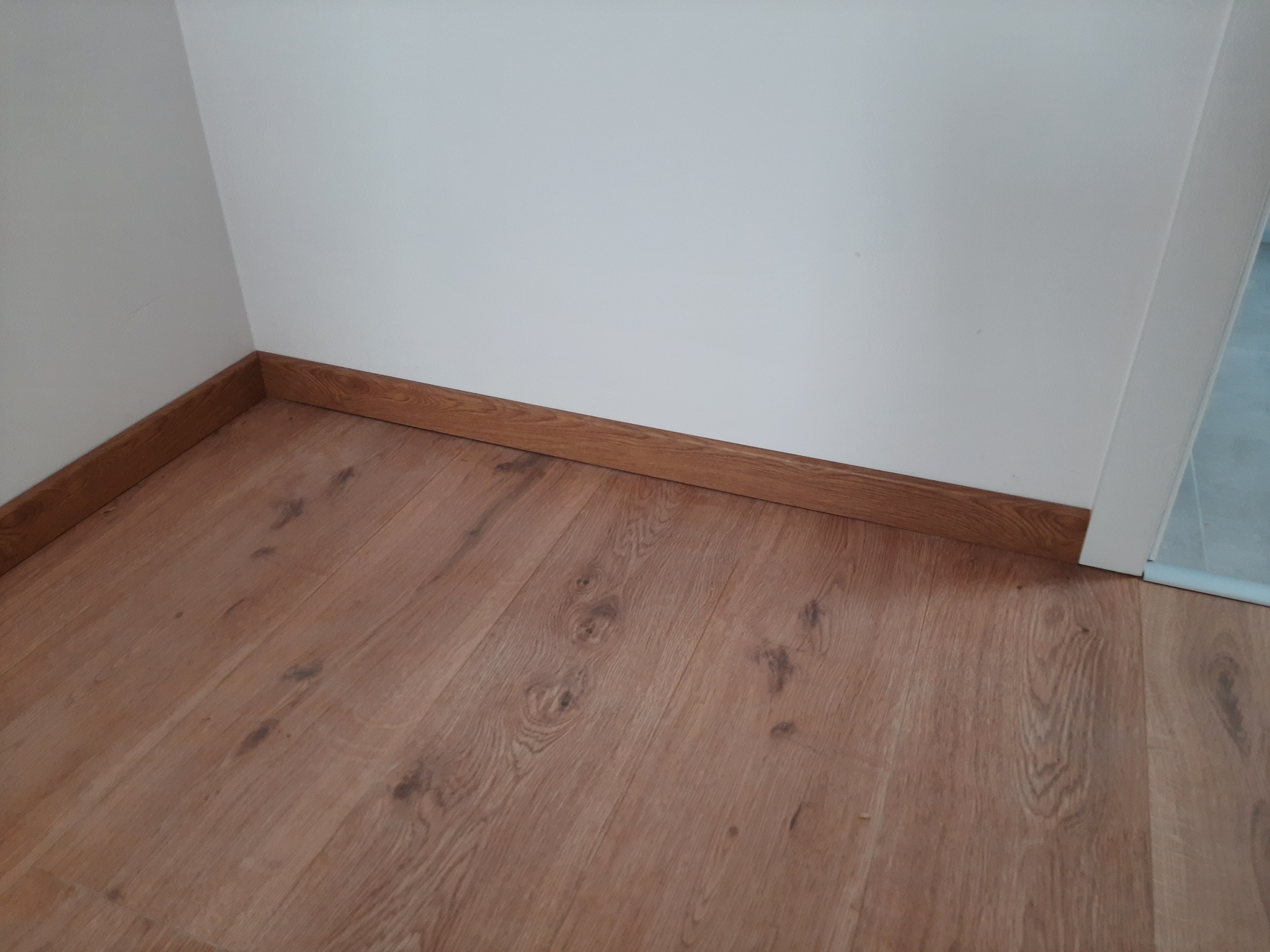 Ułożone panele laminowane Egger w rogu pokoju, widoczne listwy przypodłogowe w kolorze drewna.