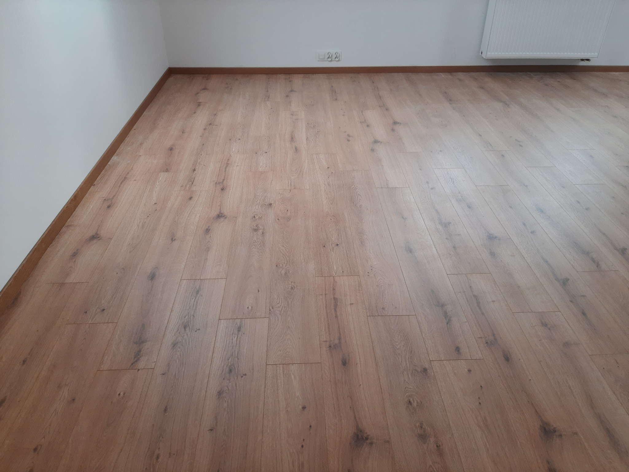 Ułożone panele laminowane Egger w pustym pokoju, widoczne gniazdka elektryczne i grzejnik na ścianie.