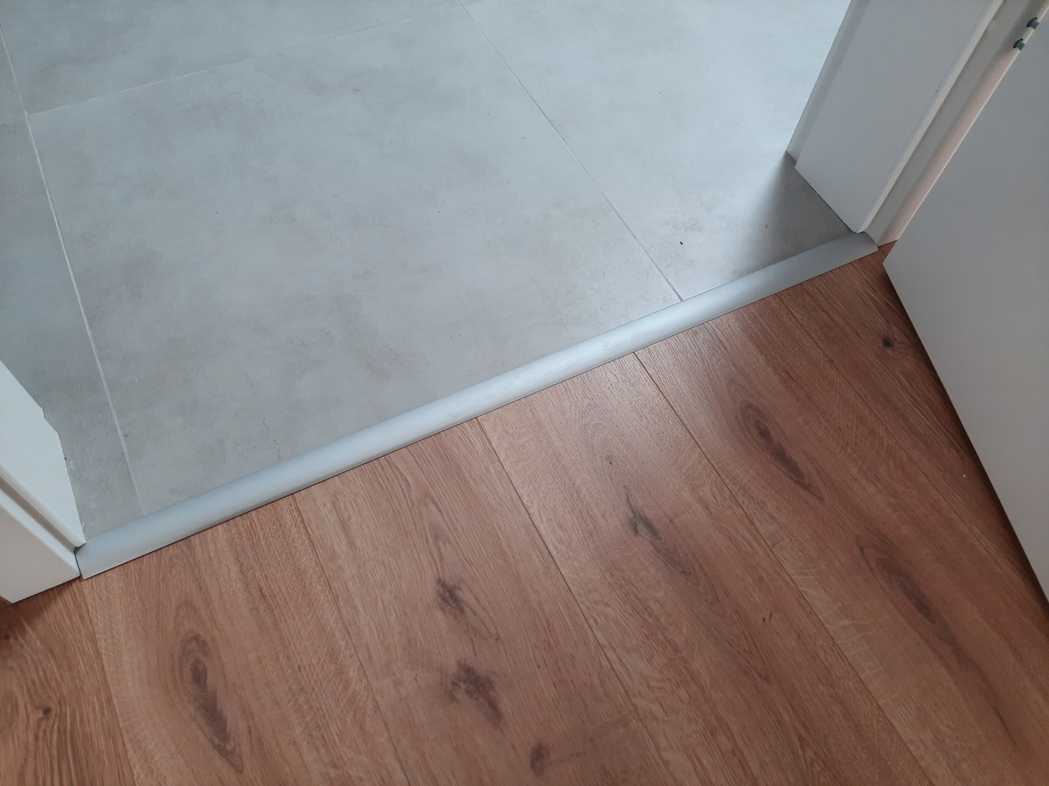 Połączenie podłogi z paneli laminowanych Egger z płytkami ceramicznymi, oddzielone szarą listwą progową przy białej futrynie drzwi.