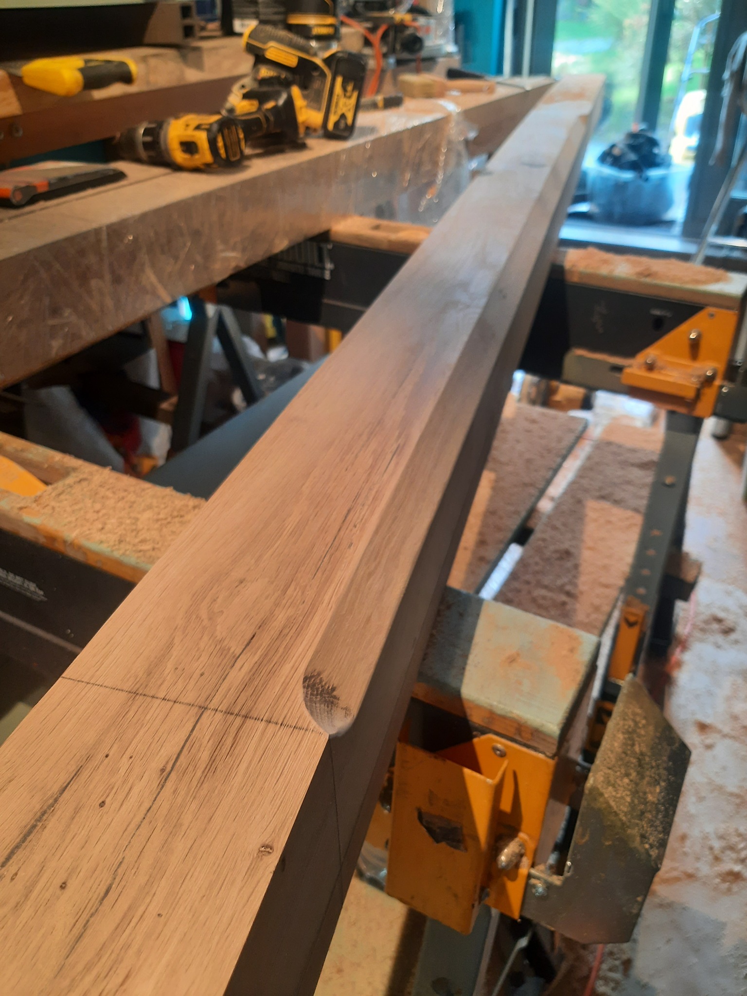 Obróbka drewnianej belki z wyfrezowanym półokrągłym wgłębieniem, widoczna na warsztatowym stole z narzędziami i wkrętarkami.