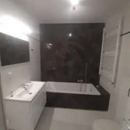 Nowoczesna łazienka z wanną obudowaną ciemnym marmurem, białą umywalką z szafką i grzejnikiem drabinkowym na ścianie w tle.