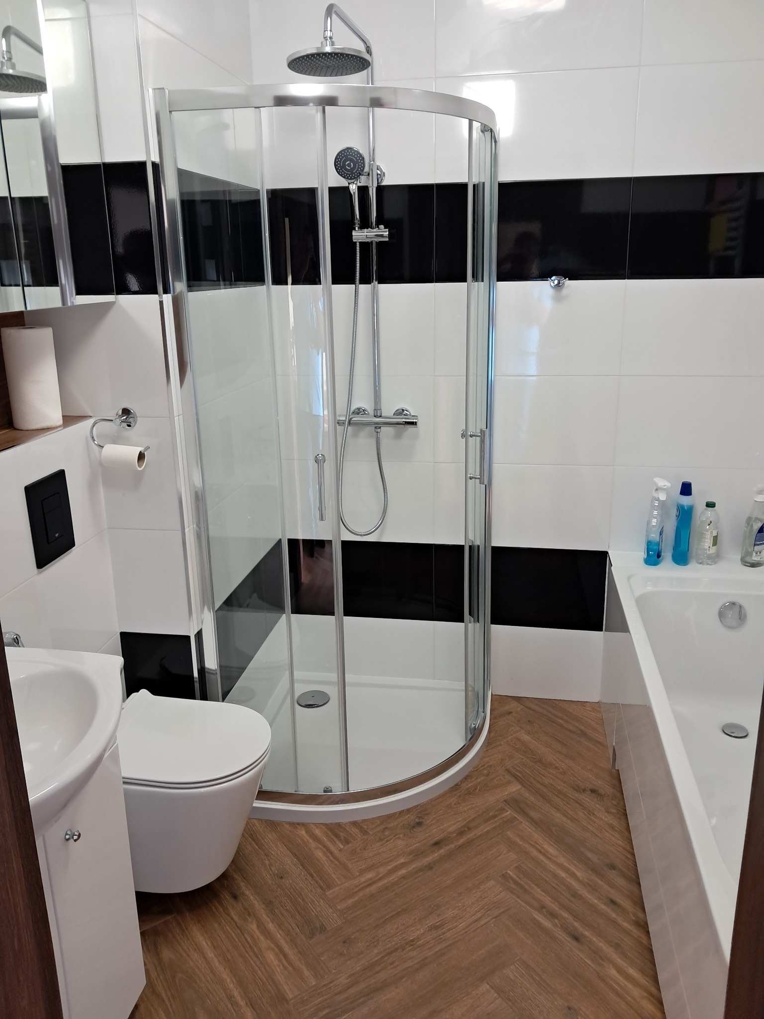 Nowoczesna łazienka z kabiną prysznicową typu walk-in, białą ceramiką, panelami ściennymi w czarno-białe pasy i podłogą z paneli imitujących drewno ułożonych w jodełkę.