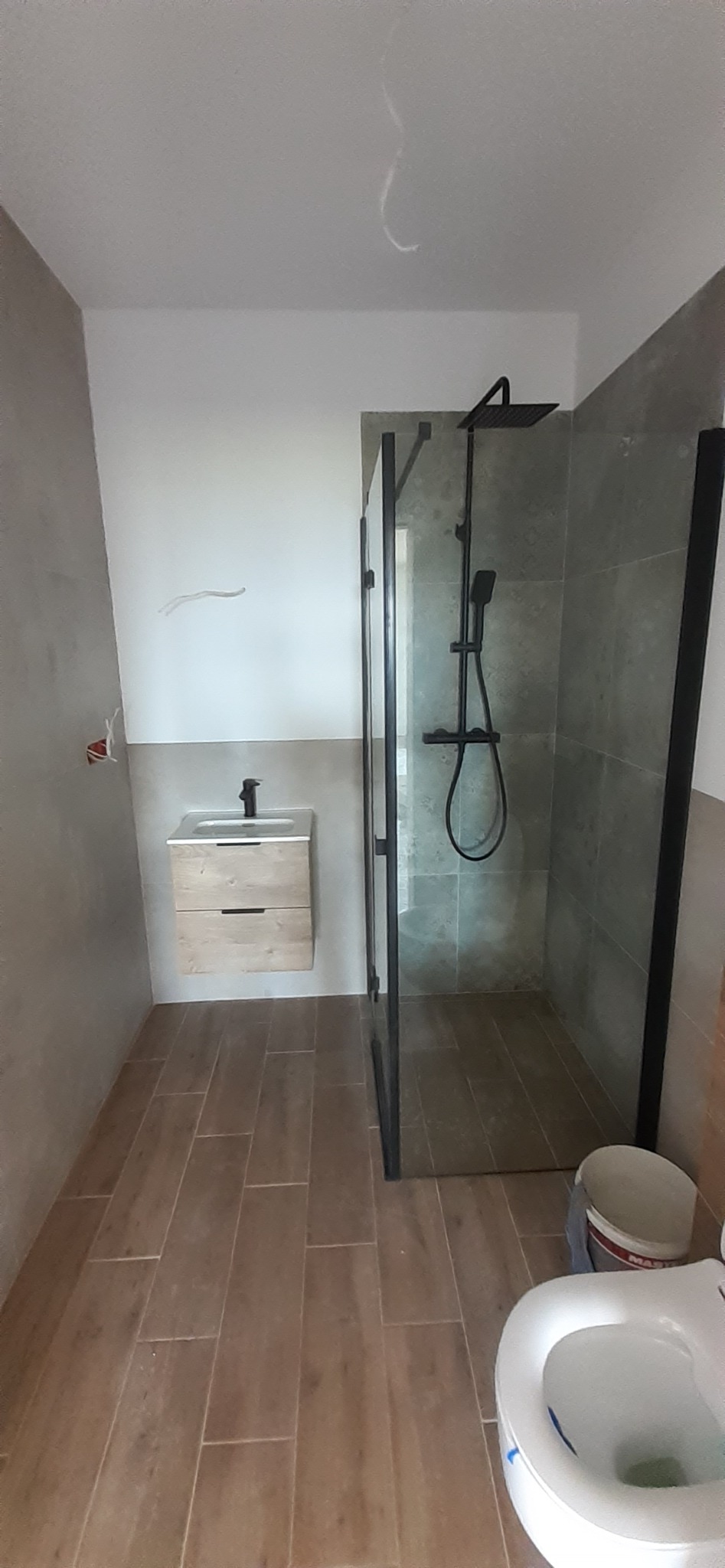 Nowoczesna łazienka w trakcie wykańczania: kabina prysznicowa typu walk-in z czarną armaturą, wisząca szafka z umywalką z drewnianymi frontami, płytki imitujące drewno na podłodze, częściowo...