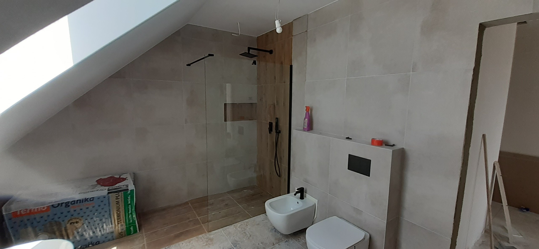 Nowoczesna łazienka na poddaszu z kabiną prysznicową typu walk-in, bidetem, toaletą i szarą płytką wielkoformatową. Widoczna paczka styropianu pod skosem dachu.