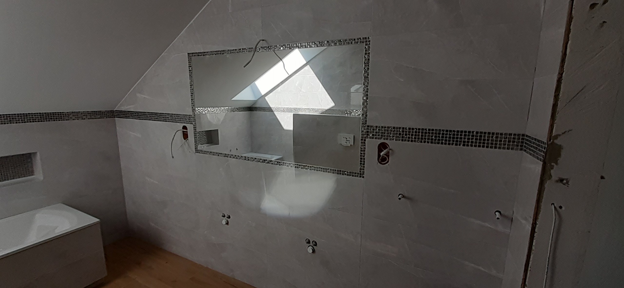 Remont łazienki na poddaszu: ściany wyłożone szarymi płytkami z dekoracyjnym pasem mozaiki, widoczne instalacje elektryczne i hydrauliczne, wnęka z oknem dachowym