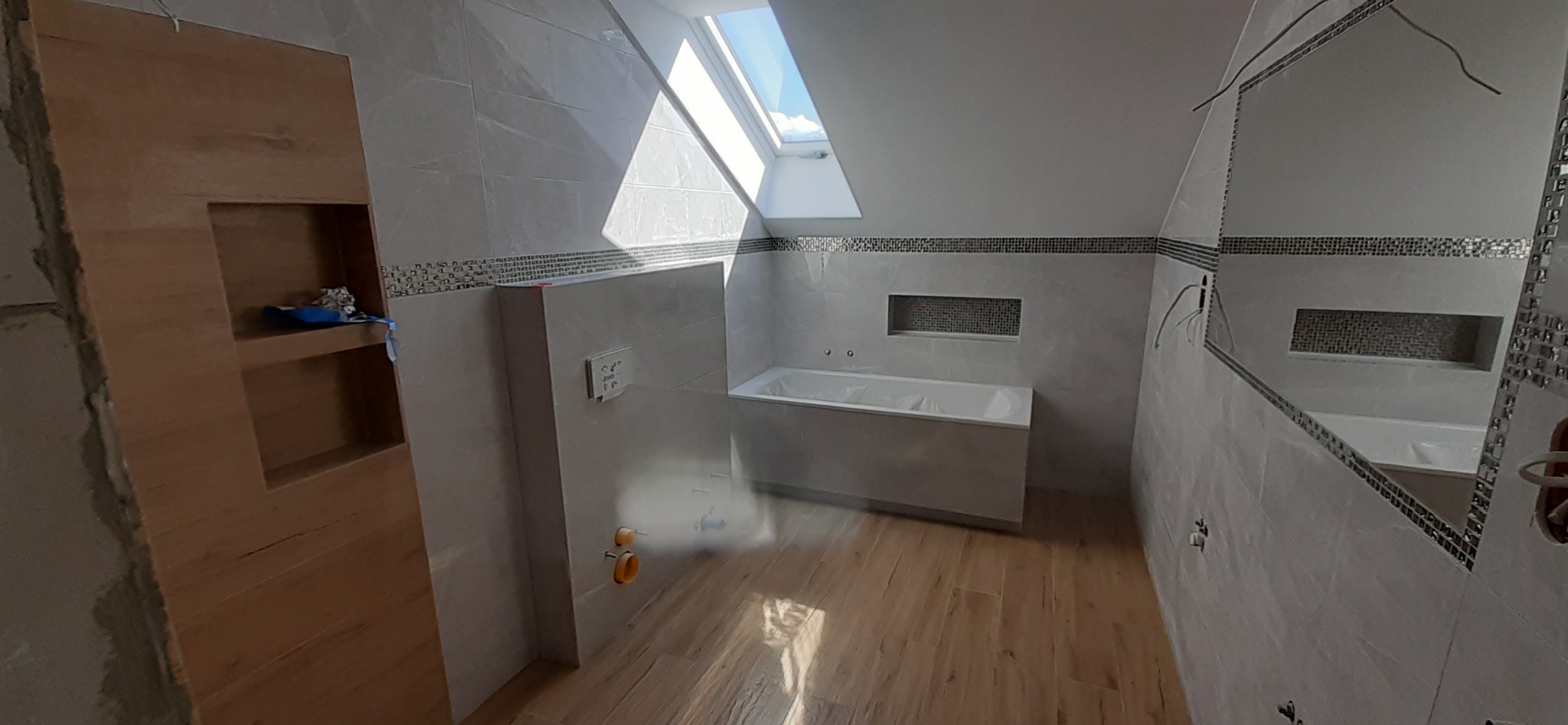 Łazienka na poddaszu w trakcie wykańczania: białe płytki, drewnopodobna podłoga, zabudowana wanna, wnęka z mozaiką, okno dachowe wpuszczające jasne światło.