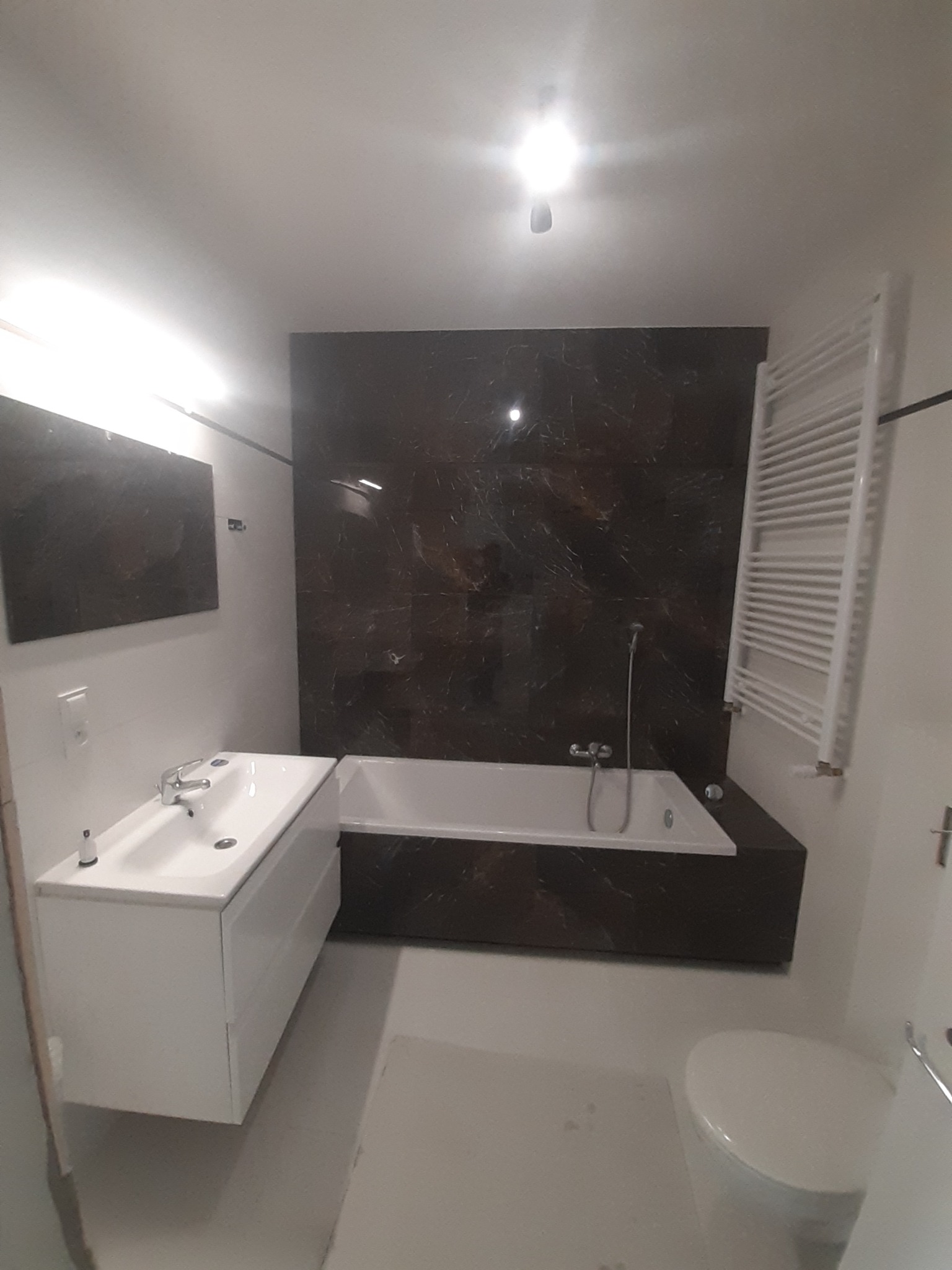 Nowoczesna łazienka z wanną obudowaną ciemnym marmurem, białą umywalką z szafką i grzejnikiem drabinkowym na ścianie w tle.