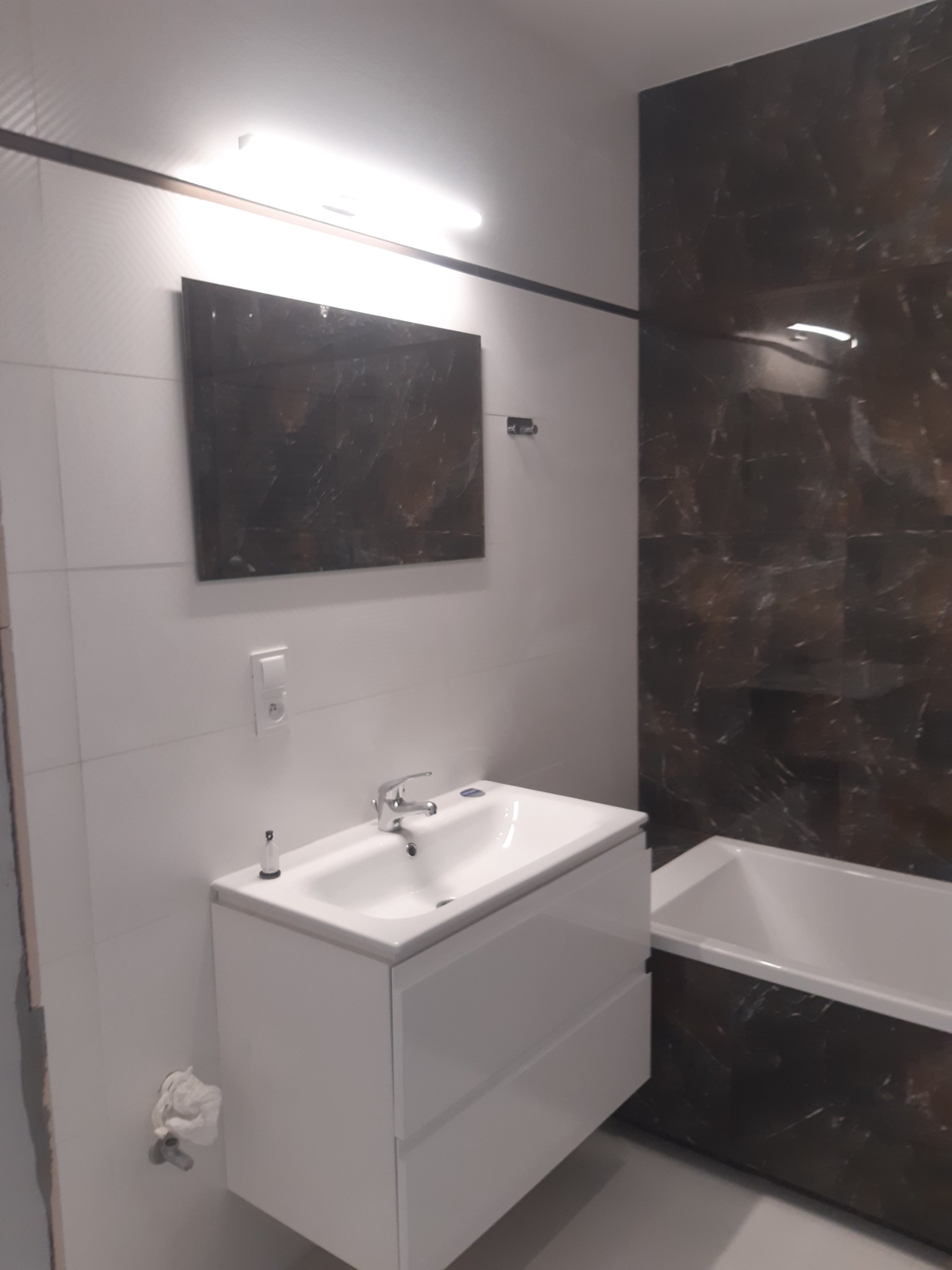 Nowoczesna łazienka z białą umywalką na szafce, lustrem w ciemnej ramie i wanną obłożoną płytkami imitującymi marmur.