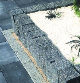 Obrzeże rabaty wykonane z kostki brukowej w kolorze grafitowym, oddzielające ciemną nawierzchnię z kostki od białego żwiru, z widocznymi roślinami w tle.