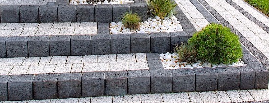 Schody z kostki brukowej w odcieniach szarości i bieli, z elementami dekoracyjnymi w postaci białych kamieni i niskich roślin ozdobnych, tworzące estetyczne przejście w ogrodzie.