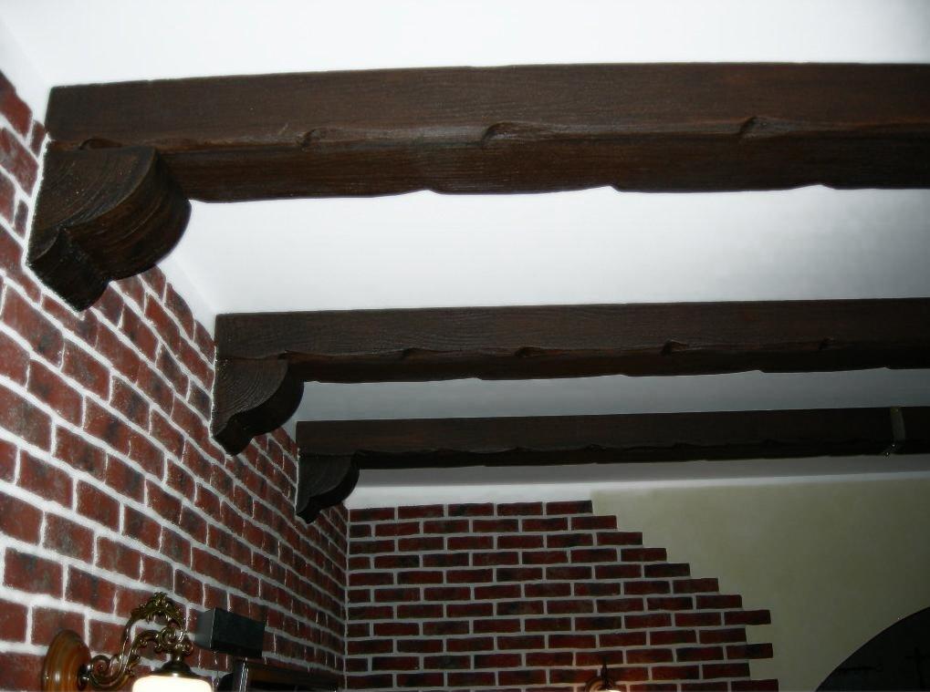 Dekoracyjne, ciemne belki drewnopodobne pod sufitem, z ozdobnymi zakończeniami, zamontowane przy ścianie z cegły klinkierowej.