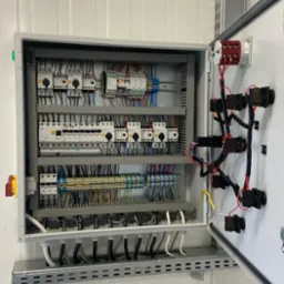 Otwarta szafa sterownicza z widocznymi elementami elektrycznymi, takimi jak przekaźniki, bezpieczniki i okablowanie, z żółto-zielonym przewodem ochronnym po prawej stronie.